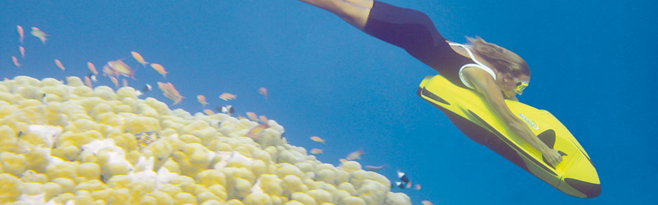 A woman riding a seabob descends past a coral bombie and a school of fish